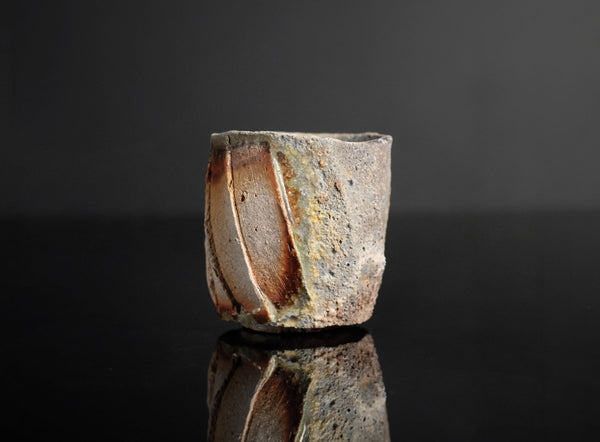A handmade ceramic guinomi with a textured, geometric design in earthy brown, beige, and white glazes, set against a dark reflective background.
 