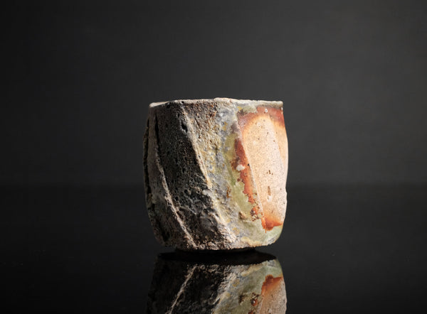 A handmade ceramic guinomi with a textured, geometric design in earthy brown, beige, and white glazes, set against a dark reflective background.