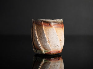 A handmade ceramic guinomi with a textured, geometric design in earthy brown, beige, and white glazes, set against a dark reflective background.