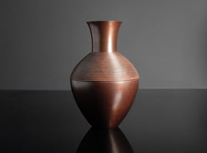 A metallic copper-colored vase with a ribbed textured pattern on the upper half, standing on a reflective black surface against a dark gray background.