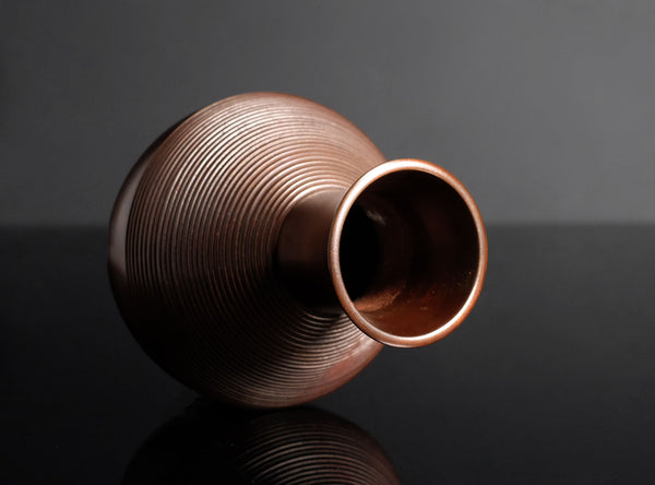 A small copper-colored vase with a ribbed texture and a wide flared opening, photographed on a dark reflective surface against a dark gray background.