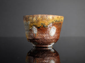 A handmade ceramic cup with a rustic, textured glaze featuring brown, gold, and white patches, displayed on a reflective black surface against a dark gray background.