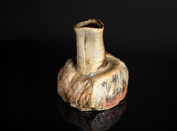 A handmade ceramic vase with a bulbous, organic-shaped base and a narrow, flared neck, featuring a textured glaze in earthy tones of beige, brown, and rust-red, set against a solid black background with a soft reflection below.