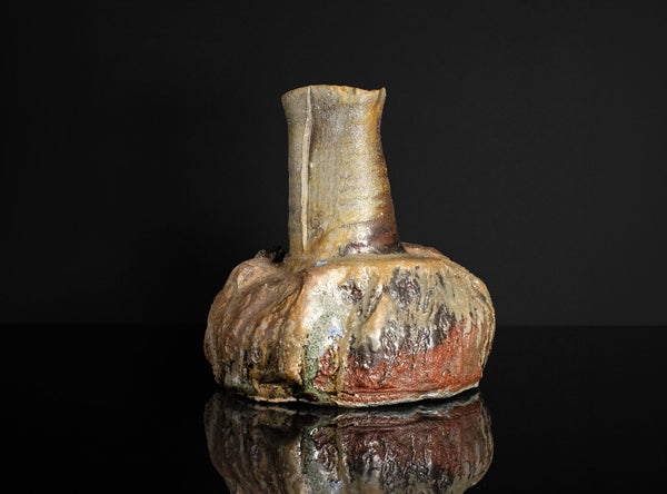 A rustic ceramic vase with a bulbous, organic base and a narrow neck, featuring a textured glaze in earthy tones of brown, beige, and reddish accents, set against a solid black background with a reflection below.