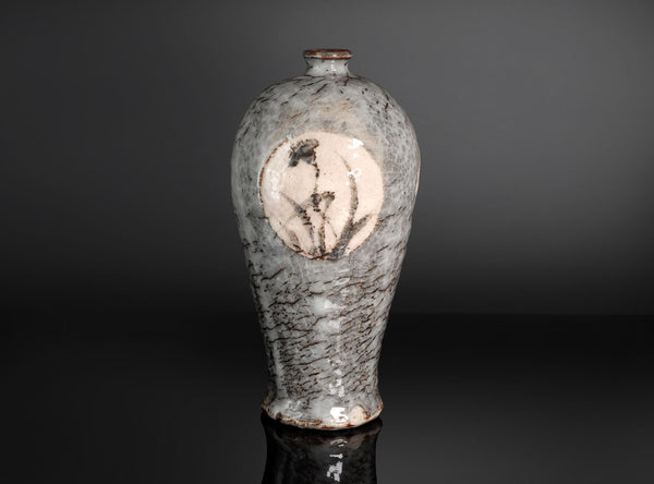 A ceramic vase with a textured gray and white glaze, featuring a circular beige inlay with a black floral illustration, set against a dark gray background.