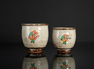 Two ceramic cups with decorative designs on a reflective surface.