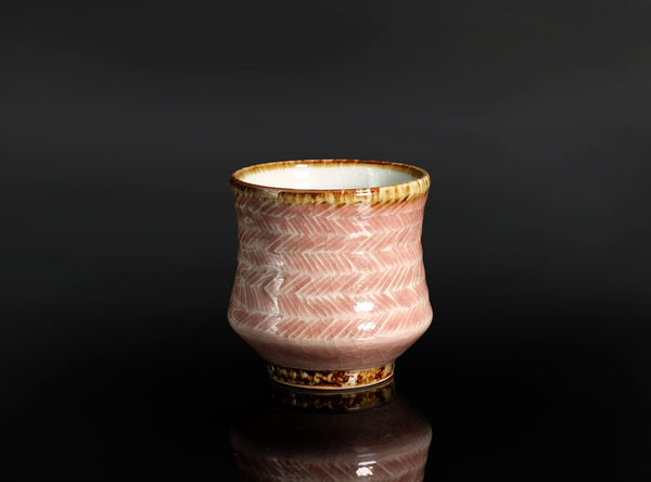 A pink ceramic tea cup with a white hand-painted herringbone pattern and a metallic gold-brown rim, set against a dark reflective background.