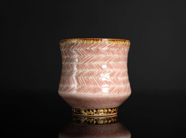 A pink ceramic tea cup with a white hand-painted herringbone pattern and a metallic gold-brown rim, set against a dark reflective background.