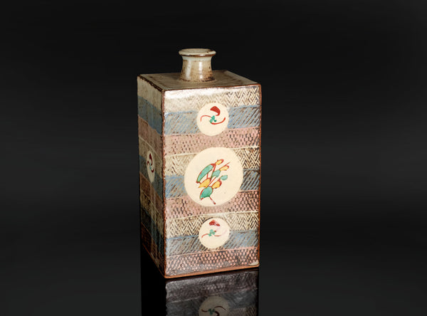 A distinctive square jar by Tatsuzo Shimaoka, combining a firm architectural form with bands of inlaid decoration and colourful floral roundels. Its cream, muted blue and pinkish-brown palette brings warmth to the straight-sided body, while the patterned surfaces reflect the artist’s inventive approach to Mashiko pottery.

The tall rectangular body rises to a broad, nearly flat shoulder and a short cylindrical neck. A gently flaring lip softens the transition between the angular vessel and its circular open