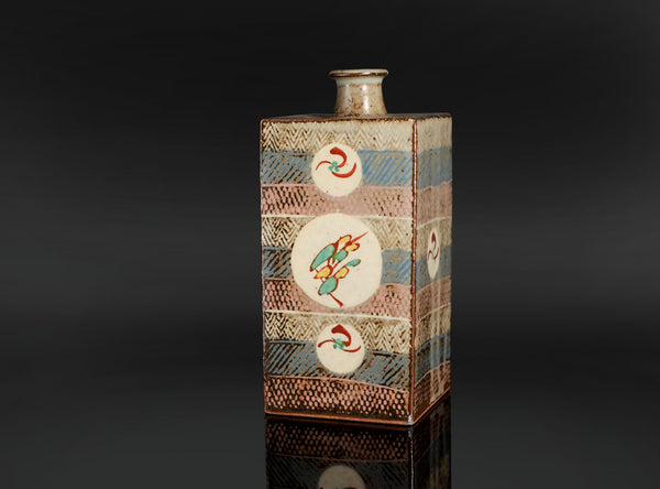 A distinctive square jar by Tatsuzo Shimaoka, combining a firm architectural form with bands of inlaid decoration and colourful floral roundels. Its cream, muted blue and pinkish-brown palette brings warmth to the straight-sided body, while the patterned surfaces reflect the artist’s inventive approach to Mashiko pottery.

The tall rectangular body rises to a broad, nearly flat shoulder and a short cylindrical neck. A gently flaring lip softens the transition between the angular vessel and its circular open