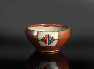 A handmade ceramic tea bowl with a glossy reddish-brown glaze, featuring a hand-painted floral motif in red, green, and black on a white patch on the front, set against a dark gray background with a reflective surface below.