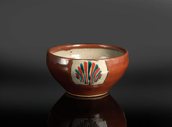 A handmade ceramic bowl with a glossy terracotta brown finish, featuring a hand-painted decorative motif on the front with red, green, and blue lines on a white background, set against a dark gray gradient background.