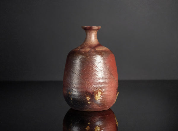 A rustic handmade ceramic bottle with a rounded body, short neck, and a glossy reddish-brown glaze with dark patches, set against a dark gray background with a reflection below.