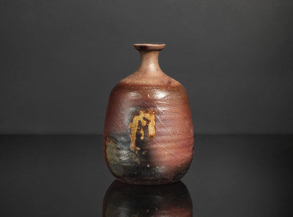 A rustic handmade ceramic bottle with a rounded body, short neck, and a glossy reddish-brown glaze with dark patches, set against a dark gray background with a reflection below.