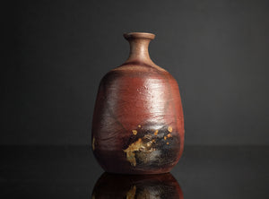 A rustic handmade ceramic bottle with a rounded body, short neck, and a glossy reddish-brown glaze with dark patches, set against a dark gray background with a reflection below.
