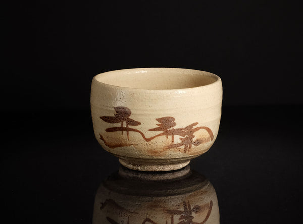 A handmade ceramic tea bowl with a textured beige glaze and brown painted brushstrokes depicting a landscape, set against a solid black background with a soft reflection below.
