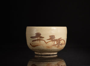 A handmade ceramic tea bowl with a textured beige glaze and brown painted calligraphic characters, set against a solid black background with a soft reflection below.
