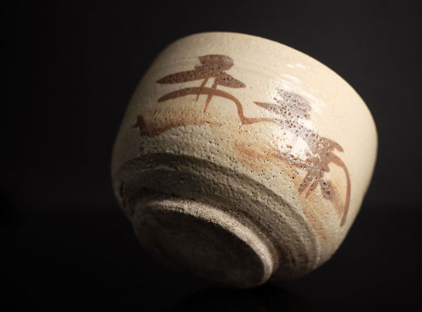 A handmade ceramic tea bowl with a textured cream glaze and brown painted calligraphic motifs, shown on a dark black background.