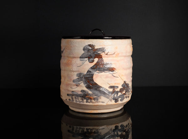 A handmade ceramic canister with a lid, featuring a textured beige body decorated with a black and brown painted motif, set against a solid black background with a reflection below.