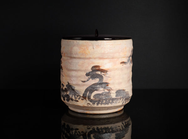 A handmade ceramic canister with a lid, featuring a textured beige body decorated with a black and brown painted motif, set against a solid black background with a reflection below.