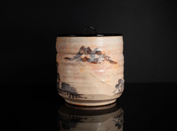A handmade ceramic canister with a lid, featuring a textured beige body decorated with a black and brown painted motif, set against a solid black background with a reflection below.
