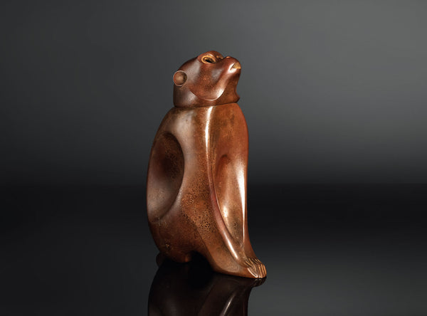 A sculptural monkey-shaped incense burner by Tsuda Eiju, distinguished by its upright seated pose and smoothly modelled brown surface. The animal raises its face, creating a lively expression while retaining the compact, self-contained outline of a small metal sculpture.

The rounded body rests on its haunches, with long forelimbs descending towards the base. A broad oval area defines the flank, introducing a shallow change in contour against the curved torso. The head is carefully simplified, with projecti