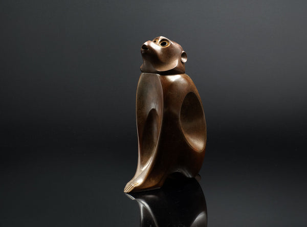 A sculptural monkey-shaped incense burner by Tsuda Eiju, distinguished by its upright seated pose and smoothly modelled brown surface. The animal raises its face, creating a lively expression while retaining the compact, self-contained outline of a small metal sculpture.

The rounded body rests on its haunches, with long forelimbs descending towards the base. A broad oval area defines the flank, introducing a shallow change in contour against the curved torso. The head is carefully simplified, with projecti