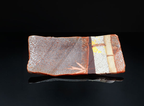 A rectangular ceramic plate with a textured orange-brown glaze, featuring a vertical band of white speckled glaze, a tan bamboo motif, and a small yellow accent, displayed on a reflective black surface.