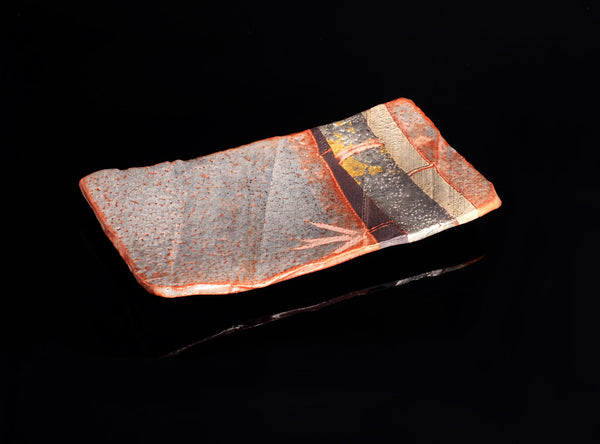 A rectangular ceramic plate with a textured, speckled glaze in gray, orange, and black, featuring a glossy glazed section on one end, set against a solid black background.