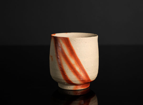 A handmade ceramic tea cup with a textured beige clay body and diagonal reddish-brown glaze stripes, set against a dark gray background with a reflective black surface below.