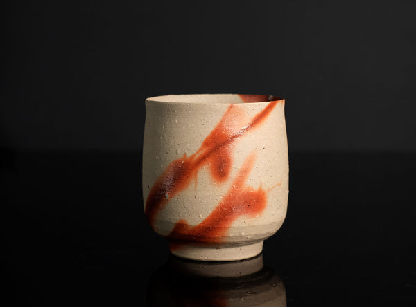 A handmade ceramic tea cup with a textured beige clay body and diagonal reddish-brown glaze stripes, set against a dark gray background with a reflective black surface below.