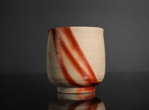 A handmade ceramic tea cup with a textured beige clay body and diagonal reddish-brown glaze stripes, set against a dark gray background with a reflective black surface below.