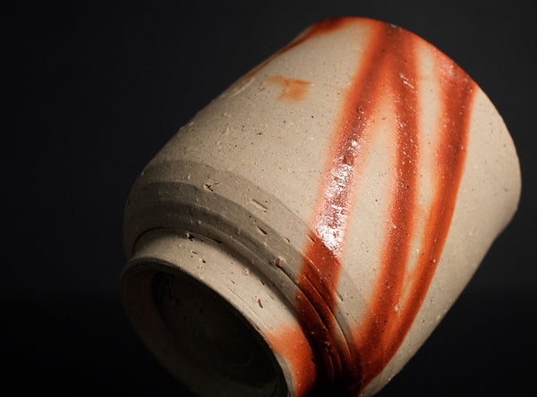 A close-up of a handmade ceramic tea cup with a textured beige clay body and diagonal reddish-brown glaze stripes, set against a dark gray background with a reflective black surface below.