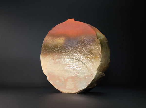 A round, textured stone or ceramic sculpture with a gradient of orange, brown, and cream, standing on a dark surface against a black background.