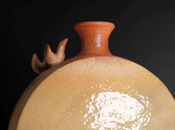 Close-up of a ceramic jug with a rounded body, narrow neck, and three small bird-like handles on the shoulder, shown against a dark background.