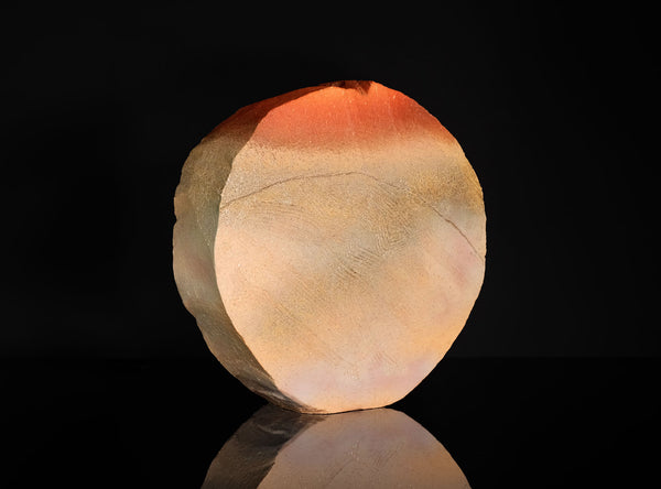 A polished natural stone sculpture with a hagi ensai flattened vase, disc-like shape, featuring a color gradient from warm orange-red at the top to light beige and cream at the bottom, displayed on a reflective black surface against a solid black background