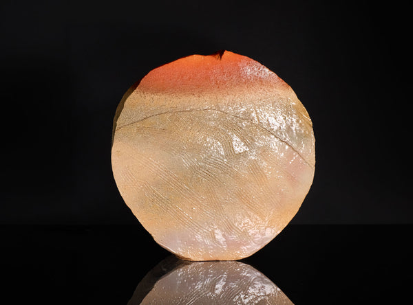 A polished natural stone sculpture with a hagi ensai flattened vase, disc-like shape, featuring a color gradient from warm orange-red at the top to light beige and cream at the bottom, displayed on a reflective black surface against a solid black background.