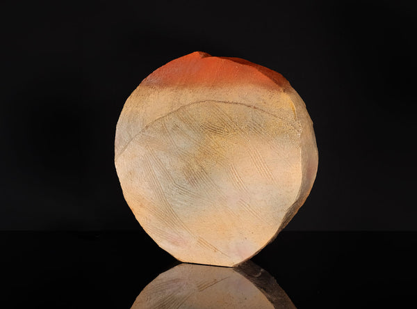 A polished natural stone sculpture with a hagi ensai flattened vase, disc-like shape, featuring a color gradient from warm orange-red at the top to light beige and cream at the bottom, displayed on a reflective black surface against a solid black background.