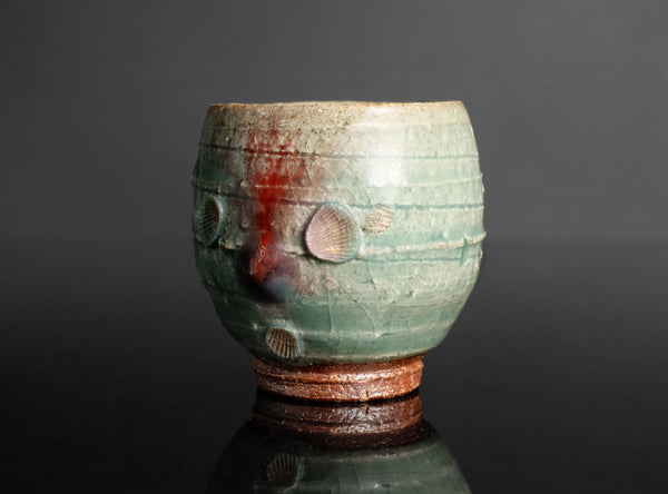 A handmade ceramic tea cup with a textured, ribbed surface and small shell inlays, featuring a green glaze with reddish-brown accents, displayed on a dark reflective surface.