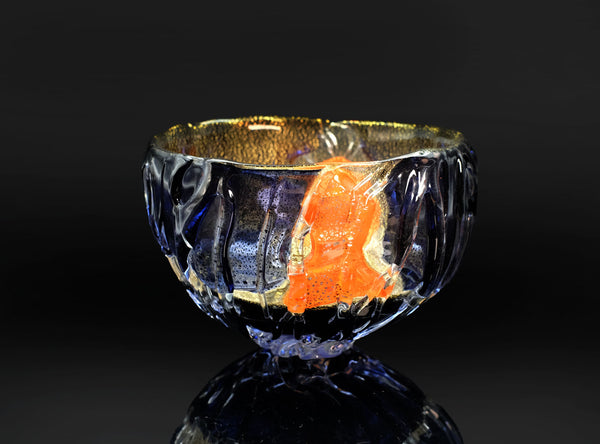A clear glass bowl with a textured, ribbed exterior and a metallic gold interior, featuring a prominent orange inlay or reflection on the front, set against a dark reflective background.