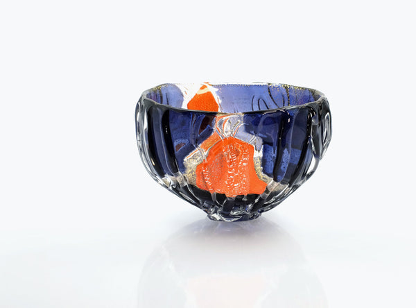 A handcrafted glass bowl with a deep blue exterior, a vibrant orange interior, and textured black and white marbled patterns, set against a clean white background with a soft reflection.