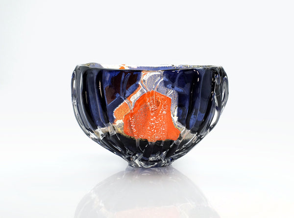 A handmade glass bowl with a deep blue exterior, featuring a prominent textured orange inlay and silver flecks, set against a clean white background with a soft reflection below.