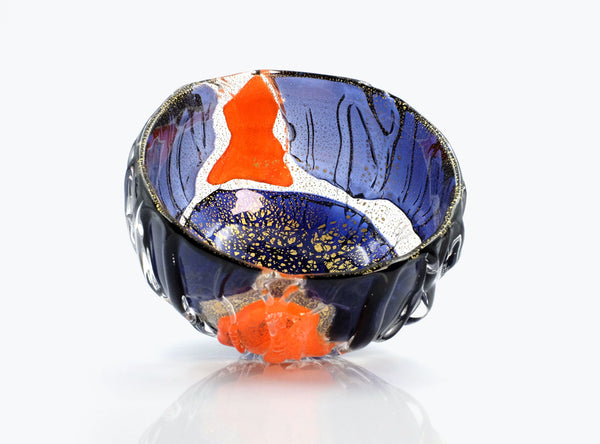 A handcrafted glass bowl with a deep blue exterior, featuring an orange and white abstract pattern on the interior and a textured, bubble-like rim, set against a clean white background with a soft reflection.