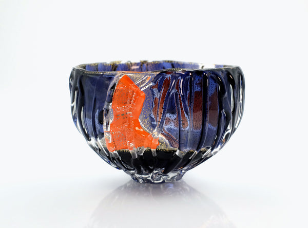 A handmade glass bowl with a deep blue and black textured exterior and a vibrant orange accent piece inside, photographed on a reflective white surface against a white background.