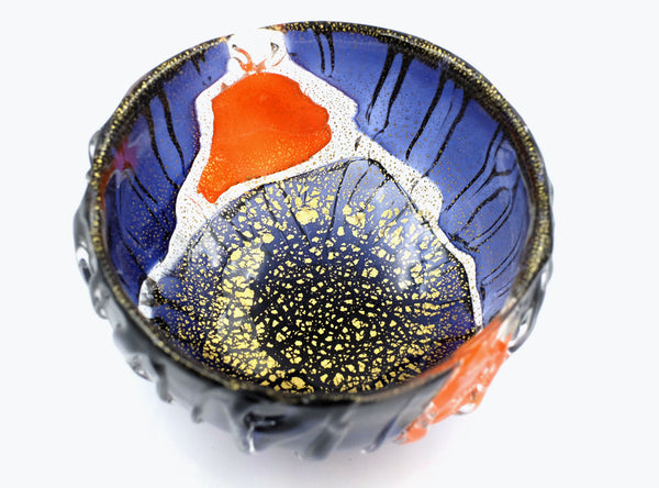 A handmade ceramic bowl with a textured, organic shape, featuring a glossy blue interior with gold leaf speckles, a white mountain motif, and a bright orange accent, set against a plain white background.